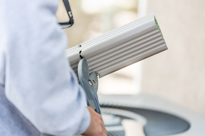 A person wearing a light blue shirt holds a set of large tin snips and cuts off a portion of a grey-colored downspout, preparing it to connect with the rain barrel in the background.