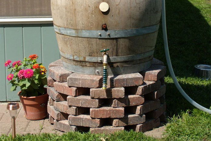 A wooden rain barrel sits atop a round stacked-brick base against the side of a blue house. The rain barrel features a spigot and a hose running from the side. Next to the barrel is a terracotta pot of pink and orange dahlias and a solar light.