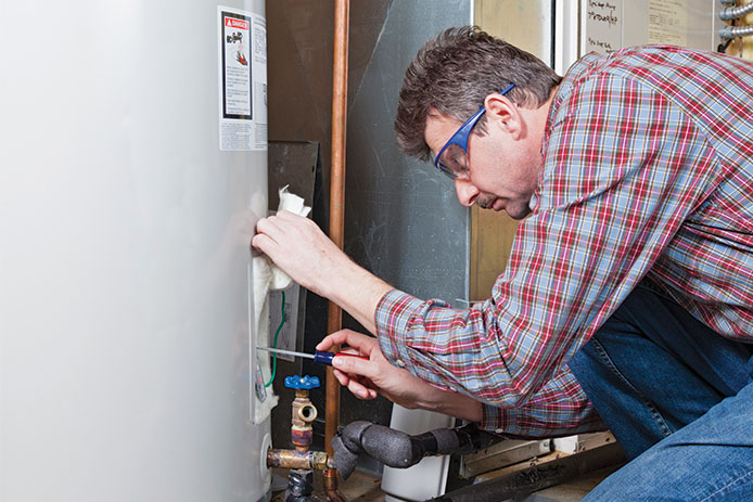 A man in a plaid shirt wearing protective glasses is working on the panel of his water heater. The water heater is light grey, and a copper pipe is running in the background.