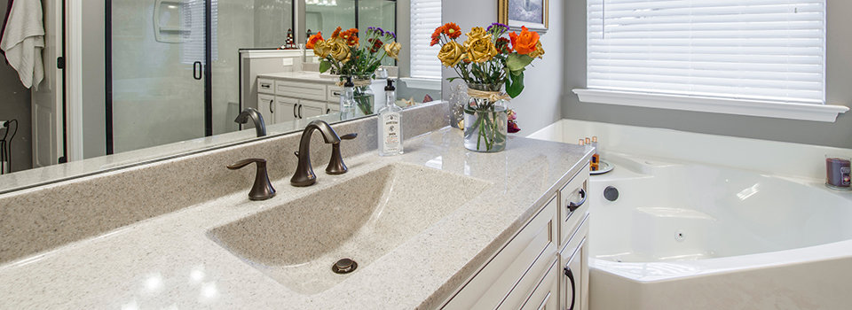 A cream-colored bathroom sink with pewter fixtures is shown alongside a bathtub. A mirror hangs in the background. Orange and yellow flowers and a soap dispenser sit atop the bathroom counter in this cream and grey, neutral-colored bathroom.