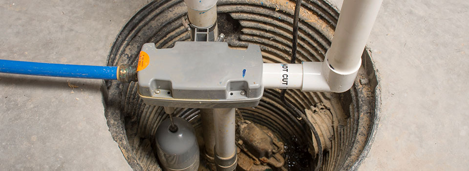 An overhead view of a sump pump in a concrete basement floor. The pump features a grey power unit, white PVC pipe running from one side, and a blue hose coming from the other.