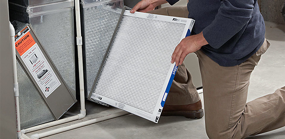 A person in khaki pants and a blue shirt is changing a white, square furnace filter.